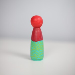 May include: A hand-painted wooden peg doll. It has a red head and torso, a turquoise base with a yellow zigzag pattern, and a red torso. The doll is standing upright.