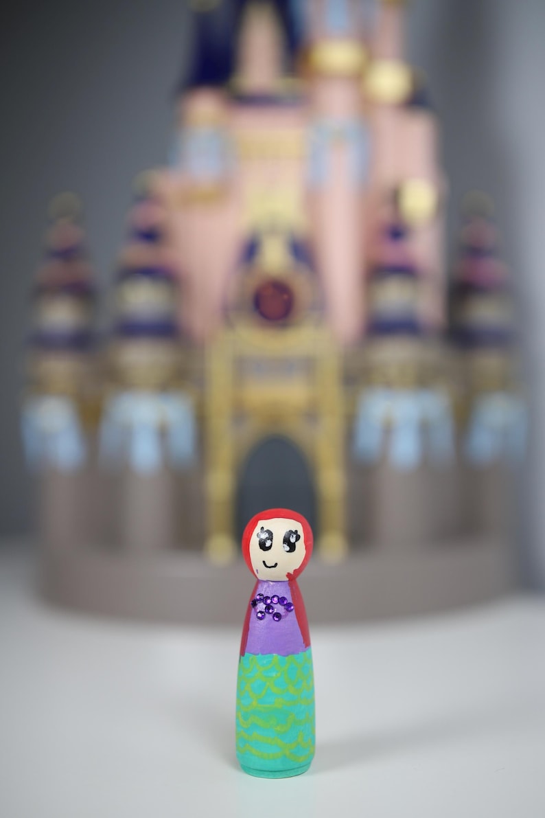 May include: A hand-painted wooden peg doll with a red hood, purple top, and turquoise skirt with a green pattern. The doll has a smiling face and is in front of a blurred pink and blue castle.