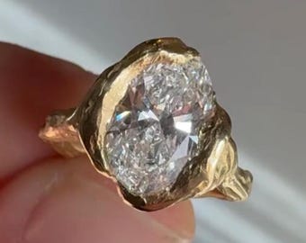 May include: A gold ring featuring a large, oval-cut clear gemstone set within a thick, textured, organic-shaped metal band. The setting has a hammered, uneven finish that surrounds the central stone, creating a bold and substantial appearance.