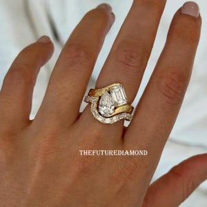 Op de afbeelding: Een gouden en diamanten ring set met een peervormige diamant als middensteen, geflankeerd door smaragd- en baguette-diamanten. De ring heeft een uniek, gebogen ontwerp met de tekst "THEFUTUREDIAMOND".