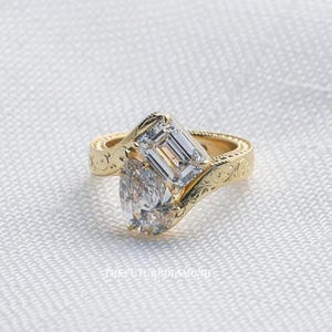Op de afbeelding: Een gouden ring met twee heldere edelstenen. E&eacute;n is peervormig, de andere smaragdgeslepen. De ringband heeft een gegraveerd bloemmotief. De ring staat tegen een witte achtergrond.
