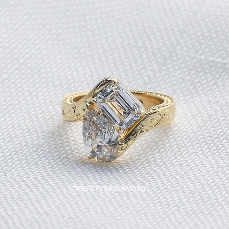 Op de afbeelding: Een gouden ring met twee heldere edelstenen. E&eacute;n is peervormig, de andere smaragdgeslepen. De ringband heeft een gegraveerd bloemmotief. De ring staat tegen een witte achtergrond.