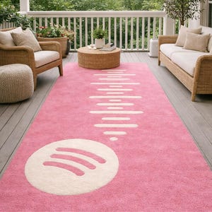 Op de afbeelding: Een roze rechthoekig tapijt met een wit Spotify-logo en golfpatroon. Het tapijt ligt op een houten terras, omringd door rotan meubels en groen, waardoor een gezellige buitenruimte ontstaat.