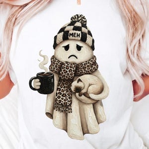 May include: A white t-shirt featuring a graphic of a sad ghost wearing a black and white checkered beanie with the text MEH, a leopard print scarf, holding a black mug with steam, and cradling a small tan cat against its body.