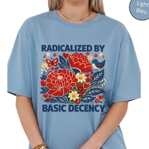Op de afbeelding: Lichtblauw T-shirt met een bloemenontwerp en de tekst "RADICALIZED BY BASIC DECENCY". De afbeelding bevat rode bloemen, vlinders en een blauwe vogel. Het shirt heeft een losse pasvorm.