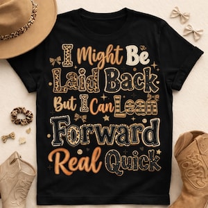 May include: Black t-shirt with the text "I Might Be Laid Back But I Can Lean Forward Real Quick" in a mix of leopard print and orange lettering. The shirt is displayed with a brown hat, scrunchie, and cowboy boots.