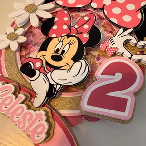 May include: A Minnie Mouse birthday cake topper with Minnie Mouse, a pink bow with white spots, and the number 2. The topper has a pink and gold glitter frame, white daisies, and the name "Celeste".