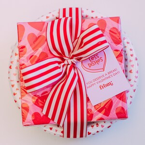 May include: A pink and red Valentine's Day gift wrapped in tissue paper with a red and white striped ribbon. The gift is tied with a tag that says "Less Dishes You Deserve a Break! Happy Valentine's Day! from:"