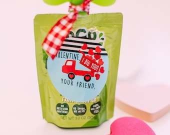 May include: A Valentine's Day gift idea featuring a red and blue truck with hearts on a blue circle tag attached to a green pouch of fruit on the go. The tag reads "Valentine, I dig you! Your friend."