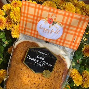 Könnte beinhalten: Eine durchsichtige Plastiktüte mit einem Kürbisgewürzkuchen darin. Der Kuchen ist mit weißem Frosting überzogen und hat ein braunes Etikett mit der Aufschrift "Marketside. Iced Pumpkin Spice Cake. Limited Time Offer". Die Tüte ist mit einem orange-weiß karierten Etikett versehen, auf dem "happy fall" mit einer weißen Kürbisillustration steht.