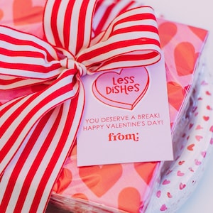 May include: A gift wrapped in red and white striped ribbon with a pink and white card that says "Less Dishes" and "You deserve a break! Happy Valentine's Day! from:". The gift is wrapped in pink napkins with red hearts.