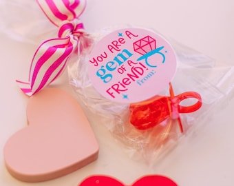 May include: A pink and white striped ribbon tied around a clear plastic bag containing a red ring pop and a pink circle sticker with the text "You are a gem of a friend!" and the words "from:" at the bottom.