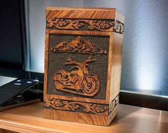 Könnte beinhalten: Holzkiste mit einem geschnitzten Motorrad-Design. Die Kiste hat einen dunklen Hintergrund mit einer Bergkette über dem Motorrad. Die Kiste ist aus hellem und dunklem Holz mit dekorativen Blättermustern gefertigt.