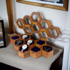 Puede incluir: Un expositor de relojes de madera con diseño de panal. La caja es de madera marrón y presenta compartimentos individuales forrados con terciopelo azul. Varios relojes se exhiben dentro de los compartimentos. La caja está sobre una superficie de color neutro.