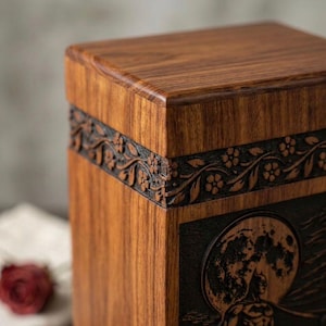 May include: A wooden box with a detailed floral design carved into a dark band around the middle. The box has a natural wood grain finish and a carved image of a full moon and a figure on the front.