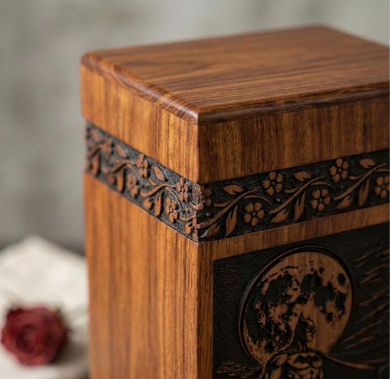 May include: A wooden box with a detailed floral design carved into a dark band around the middle. The box has a natural wood grain finish and a carved image of a full moon and a figure on the front.