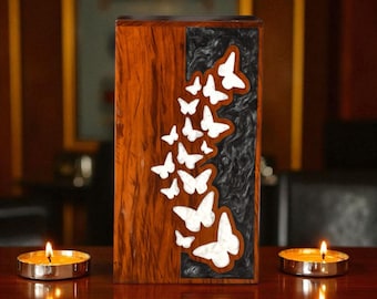 May include: A rectangular wooden box with a dark brown finish and a black textured inlay. White butterfly cutouts cascade down the side. Two lit candles sit on a wooden surface, flanking the box. The box is likely a decorative item.
