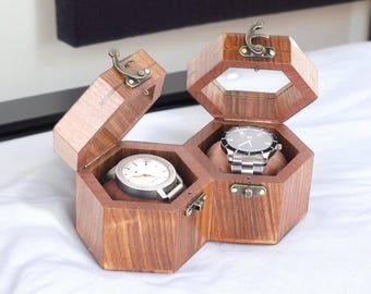 May include: A wooden watch box with two compartments, each holding a wristwatch. The box is hexagonal with a hinged lid and a clasp. The watches have silver-toned bands and faces. The box is made of a dark wood.