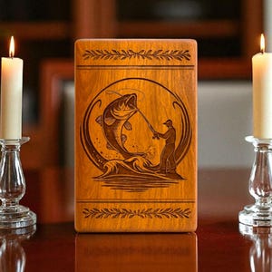 May include: A wooden box with a fishing scene engraving, flanked by two lit white candles in clear glass candle holders. The box features a circular design of a fisherman catching a fish.