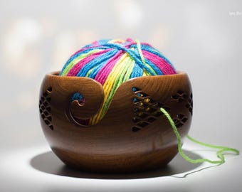 May include: A wooden yarn bowl with a vibrant ball of yarn inside. The yarn is a mix of pink, blue, yellow, and green. The bowl has a smooth, polished finish and a decorative swirl on the side. A strand of green yarn extends from the bowl.