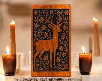 May include: A wooden box with a carved deer design, framed by floral patterns. The deer is a light brown color, set against a black background. Lit candles surround the box, creating a warm, inviting ambiance. A decorative item with text is at the bottom.