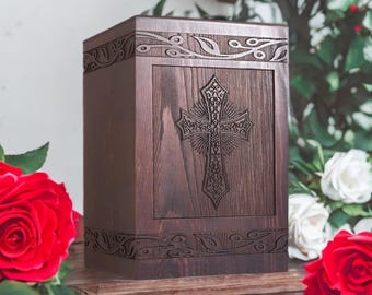 May include: A dark brown wooden urn with a carved cross design on the front panel. The urn has decorative carvings along the top and bottom edges. Red roses and white roses are in the background.