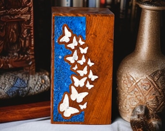 Puede incluir: Caja de madera con incrustaciones de resina azul y recortes de mariposas blancas. La caja es rectangular y de madera marrón. Las mariposas están dispuestas verticalmente. Objetos decorativos en el fondo.