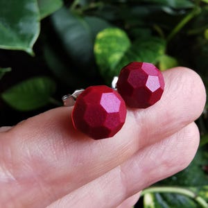May include: A pair of stud earrings featuring deep red, multifaceted, spherical beads with a subtle shimmer. The earrings are designed with silver-toned metal posts and butterfly backings, showcasing a geometric, faceted surface texture.