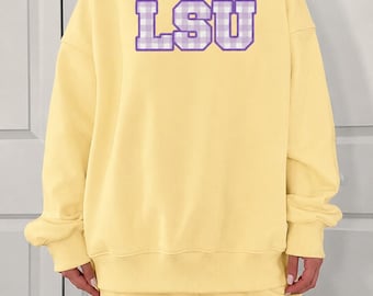 May include: A pale yellow long-sleeve sweatshirt with a mock neck collar. The center of the chest features the letters LSU in a purple and white gingham check pattern with a solid purple outline, printed in a bold, block-style collegiate font.