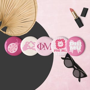 May include: A selection of pink and white circular badges with diverse designs, including a ladybird, Greek letters, and a lion. Also shown are black sunglasses, lipstick, and a woven fan.