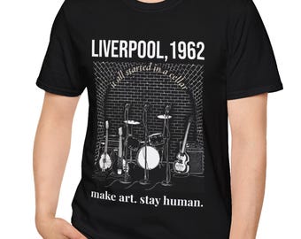 May include: Black t-shirt featuring a band graphic set in a brick cellar. The text reads "LIVERPOOL, 1962" above the image, with "it all started in a cellar" and "make art. stay human." below. The design includes guitars, drums, and an amplifier.