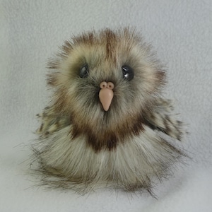 May include: A small plush owl figurine covered in long, shaggy faux fur in shades of brown, tan, and cream. The owl features two large, round, glossy black eyes and a smooth, light-coloured, triangular beak centred on its face.