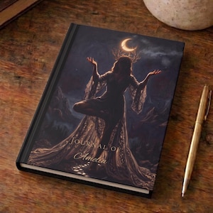 Könnte beinhalten: Ein Notizbuch mit festem Einband und einer dunklen, atmosphärischen Illustration einer Figur mit Geweih, die nach einer Mondsichel greift. Der Einband zeigt den Text "JOURNAL OF Amelia" in goldener Schrift. Ein goldfarbener Metallstift liegt daneben.