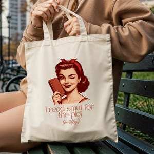 May include: Cream-colored tote bag featuring a retro illustration of a winking woman holding a book. The text "I read smut for the plot (mostly)" is printed below the image in a reddish-brown color. The bag has two handles.