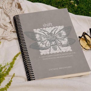 May include: A grey spiral-bound journal titled Shift, featuring a moth illustration and the text A Self-Guided Journal Based on the Principles of Neuroplasticity and Catskill Pressworks printed on the front cover in white and black ink.