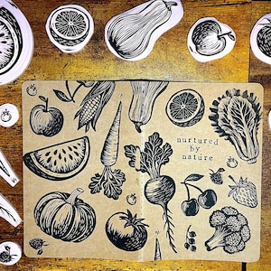 May include: Black and white linocut prints of various fruits and vegetables, including watermelon, lemon, squash, apple, and tomato. A notebook with similar illustrations and the text "nurtured by nature" is in the center. The prints are arranged on a wooden surface.