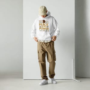 Op de afbeelding: Witte hoodie met de tekst "PEACE IS MY POWER" in bruin. Het ontwerp bevat een bruine afro-afbeelding en decoratieve patronen. De persoon draagt een kaki cargobroek en een beige baseballpet.