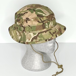 Op de afbeelding: Camouflage vissershoed met een brede rand en verstelbare kinband. De hoed heeft een mix van groene, bruine en beige kleuren. De hoed wordt gepresenteerd op een mannequinhoofd.