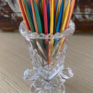 May include: A clear, cut-glass toothpick holder filled with colorful toothpicks. The toothpicks are various colors, including red, yellow, blue, green, and orange. The holder has a decorative, ornate design.
