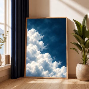 May include: A rectangular wall art print in a light wood frame featuring a high-contrast image of fluffy white cumulus clouds against a deep navy blue sky. The artwork is oriented vertically, showcasing a dense cloud formation filling the lower left.