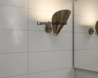 May include: A brass-colored wall sconce with a curved, fan-like shade is mounted on a white tiled wall. The sconce is lit, casting a warm glow. The text "LampfinixDecor" is visible on the wall. The sconce is a modern design.