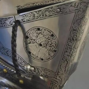 May include: Close-up of a polished silver helmet with intricate engraved floral and geometric patterns. A circular emblem features a detailed design. A black braided strap and gold rivets add contrast to the metallic surface.