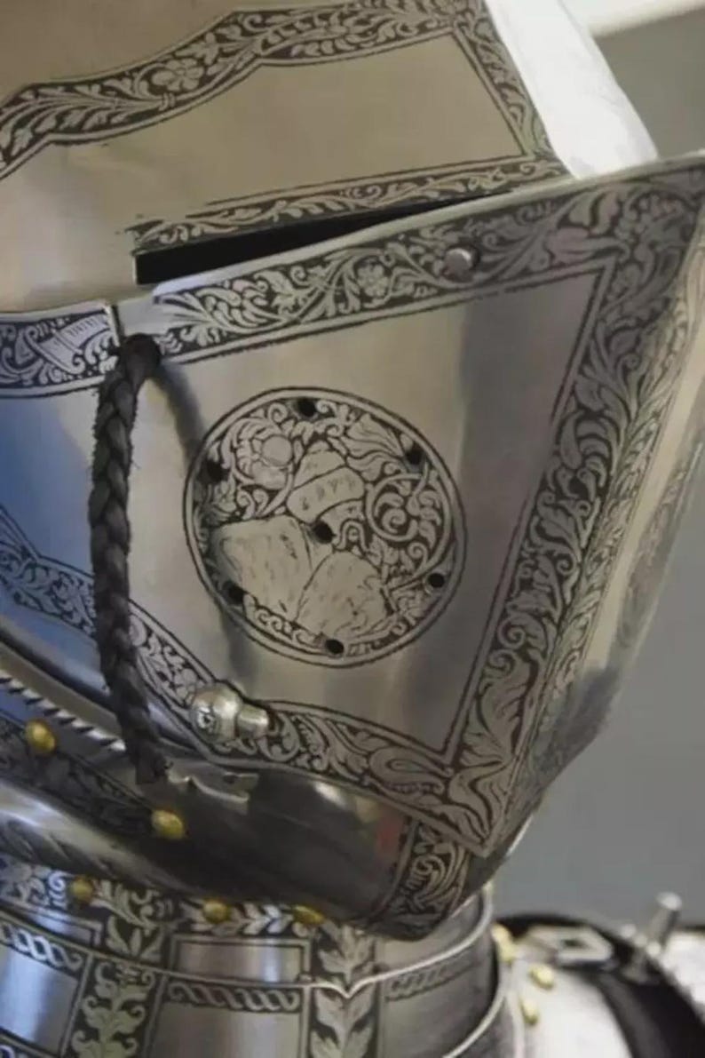 May include: Close-up of a polished silver helmet with intricate engraved floral and geometric patterns. A circular emblem features a detailed design. A black braided strap and gold rivets add contrast to the metallic surface.
