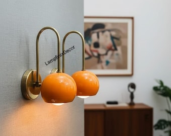 May include: Two wall-mounted sconces with a mid-century modern design. Each sconce features a round, orange globe shade and a brass-colored, curved arm. The sconces are mounted on a gray wall, providing a warm, inviting glow.