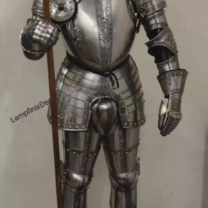 May include: Full suit of polished steel armor, including a helmet, breastplate, and leg protection. The armor is highly detailed with rivets and decorative elements. A wooden spear is held in the right hand. The armor is displayed on a wooden stand.