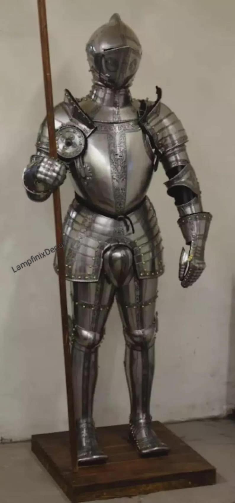 May include: Full suit of polished steel armor, including a helmet, breastplate, and leg protection. The armor is highly detailed with rivets and decorative elements. A wooden spear is held in the right hand. The armor is displayed on a wooden stand.