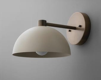 May include: A wall-mounted light fixture with a cream-colored, dome-shaped shade. The fixture has a bronze-colored arm and a circular backplate. The light bulb is visible inside the shade. This modern sconce is designed for indoor use.