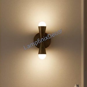 May include: A contemporary wall light with a brass finish, featuring two spherical light bulbs. The fixture has a symmetrical hourglass shape and is mounted on a circular backplate. The text "LampfinixDecor" is visible.