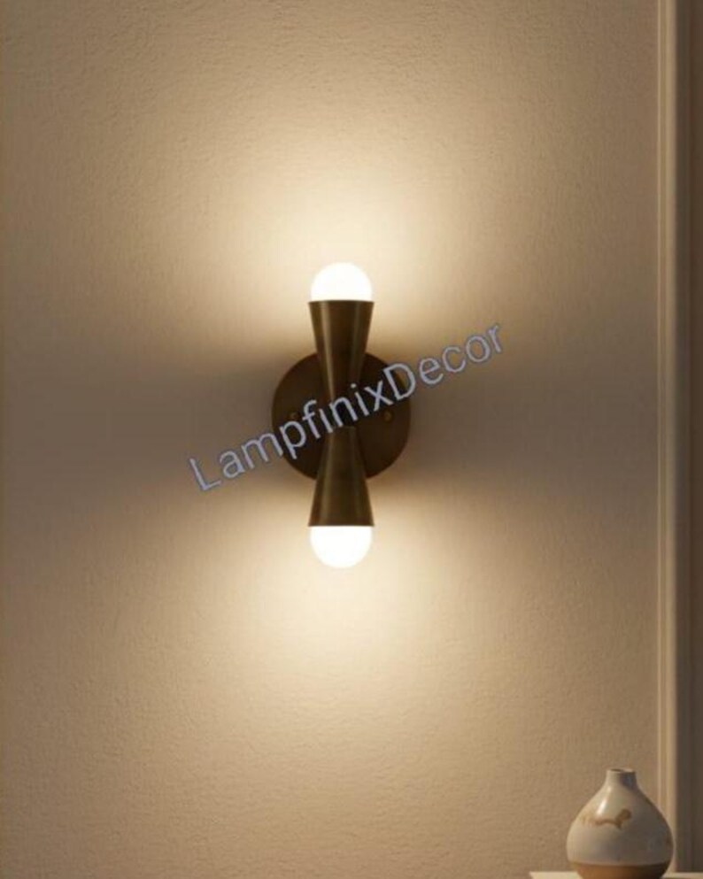 May include: A contemporary wall light with a brass finish, featuring two spherical light bulbs. The fixture has a symmetrical hourglass shape and is mounted on a circular backplate. The text "LampfinixDecor" is visible.