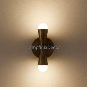 May include: A contemporary wall light with a brass finish. The fixture features two conical shades with a circular base and two white globe lights. The text "LampfinixDecor" is visible on the wall.